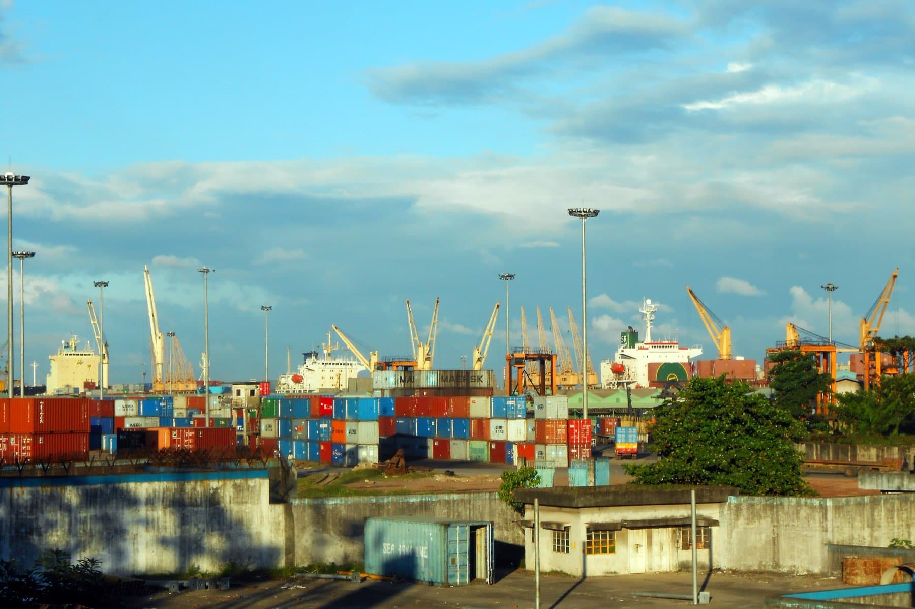 Containers stacked in a working port with ships and quay cranes behind.