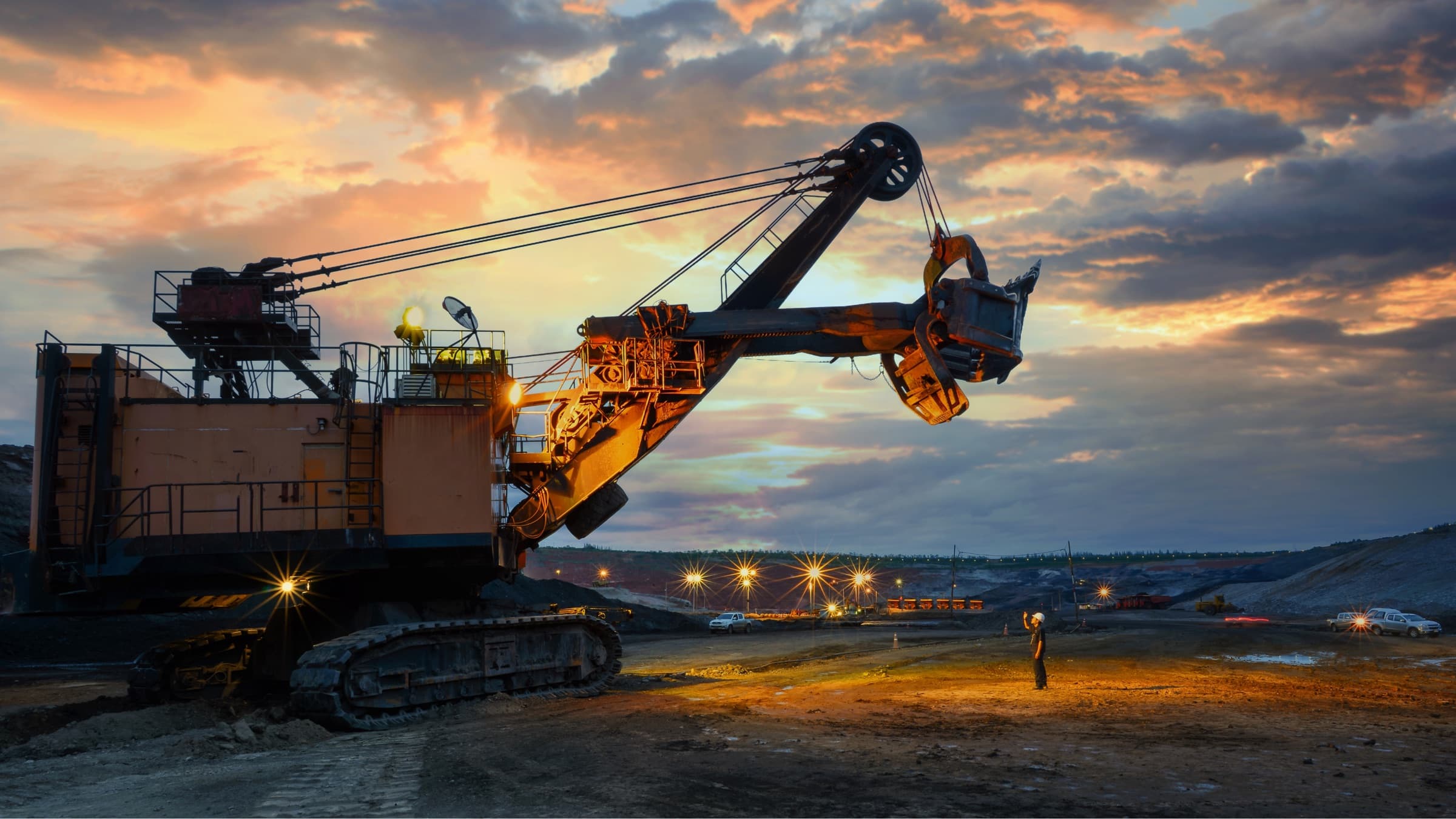 A large mining shovel standing on the floor of an open pit at dusk, a worker small in frame beside it