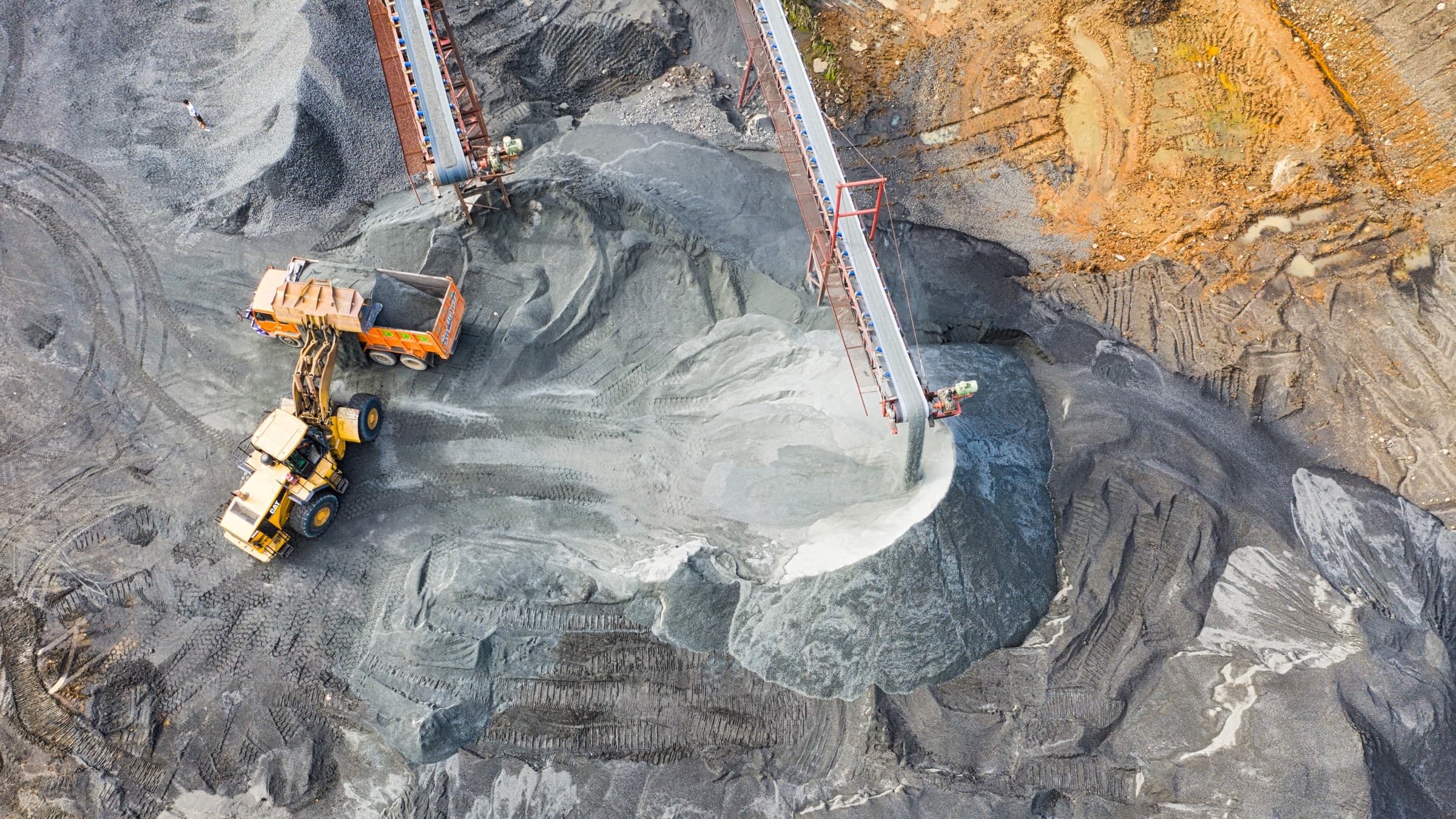 Ore stockpiles seen from above, a wheel loader and haul truck working the pile beside a conveyor